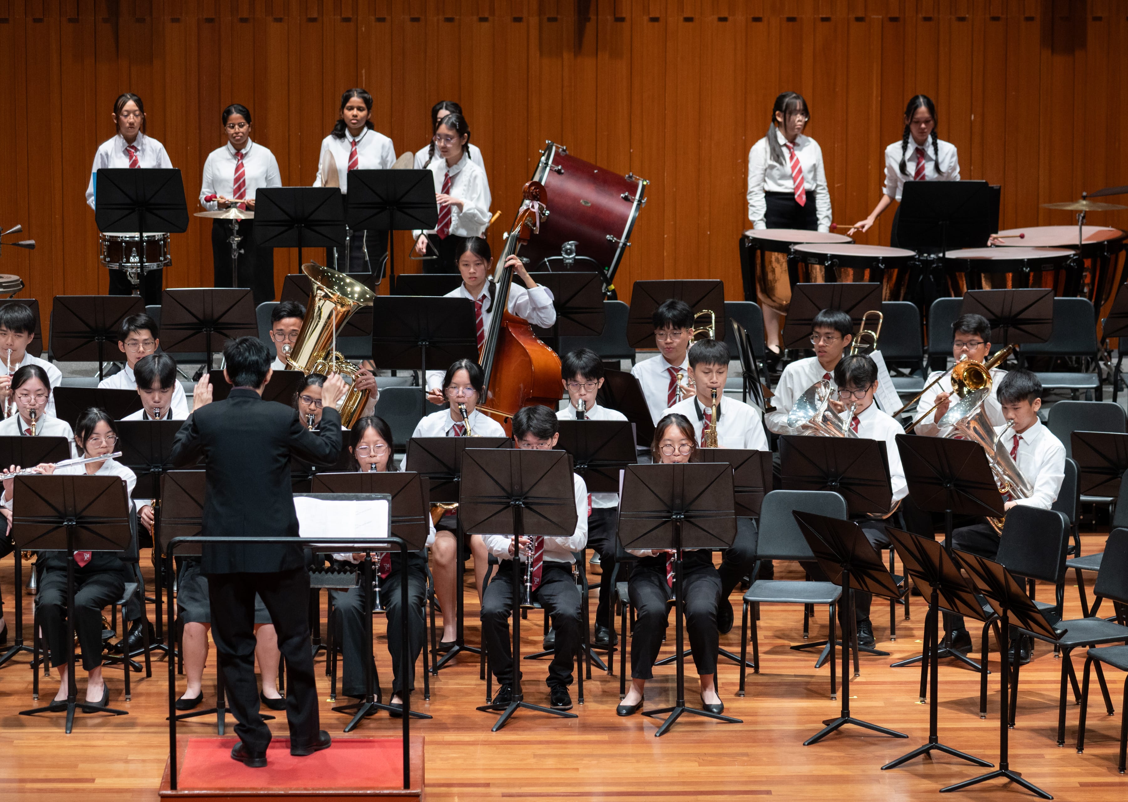 A large student symphonic band in white shirts and red ties performs on a concert hall stage, with a conductor in black facing them from the front. Musicians play a wide range of instruments including brass, woodwind, double bass, and percussion arranged across multiple rows.